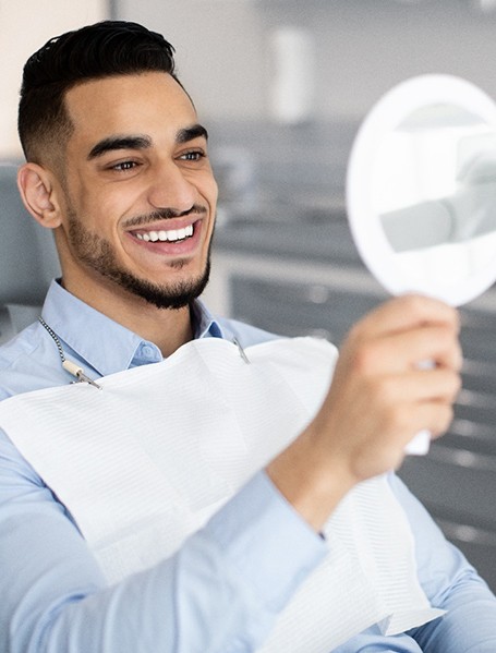 Man in dental chair smiling at handheld mirror with dentist in background
